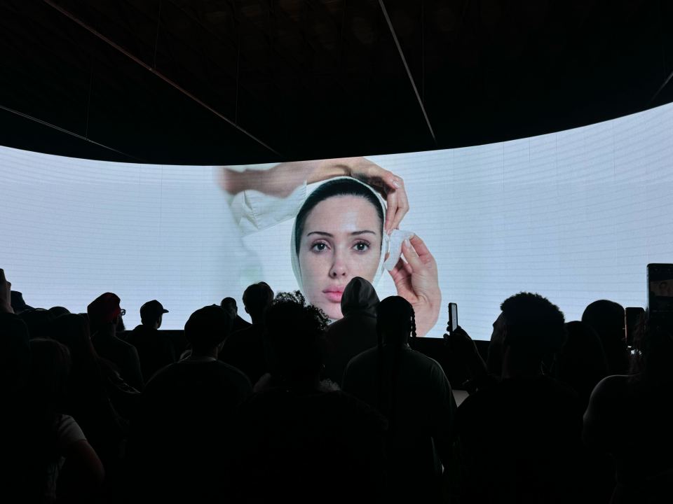 Audience watching a large screen showing a woman applying a face mask.